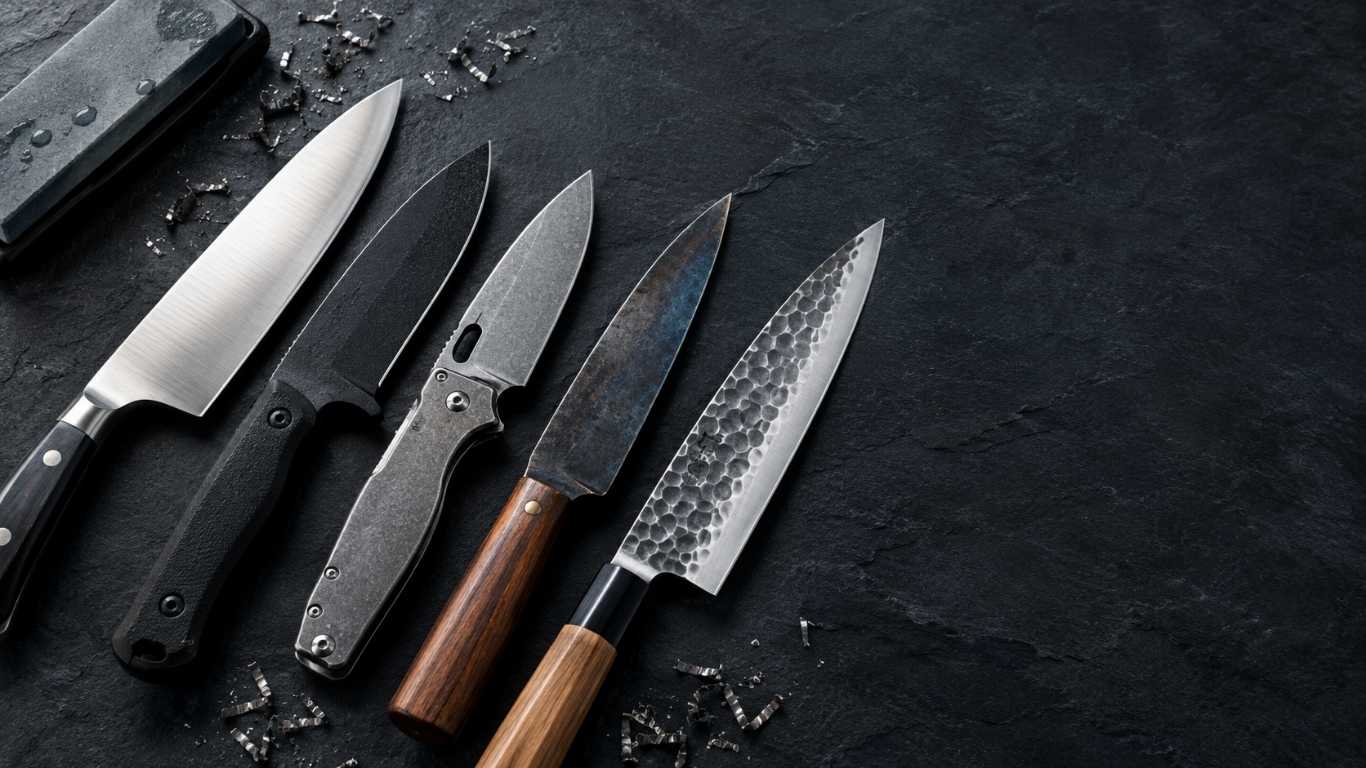 best steel for knives feature , Five knife blades in different steels fanned out on a slate surface, from kitchen chef's knife to outdoor fixed blade